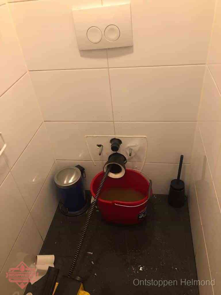 Een ontstopte wand-wc met verwijderde toiletpot waar een ontstoppingsveer in de wanduitgang verdwijnt, het achterhalen waar de verstopping zit in Helmond