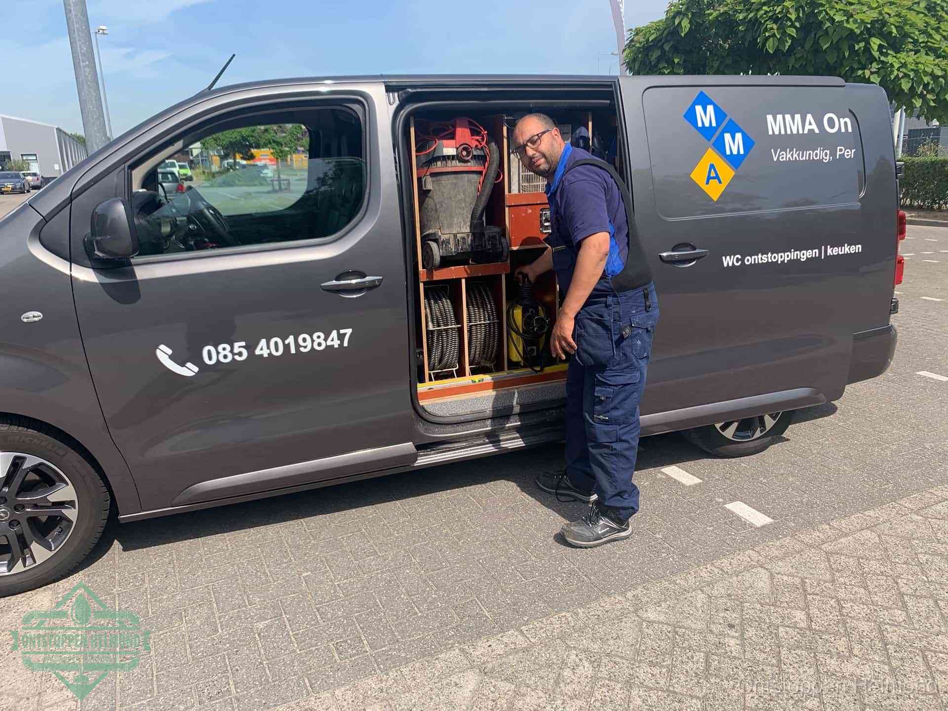 Een monteur bij de open schuifdeur van zijn bedrijfsbus op een parkeerplaats, klaar om de oorzaak van terugkerende afvoerverstoppingen aan te pakken in Helmond
