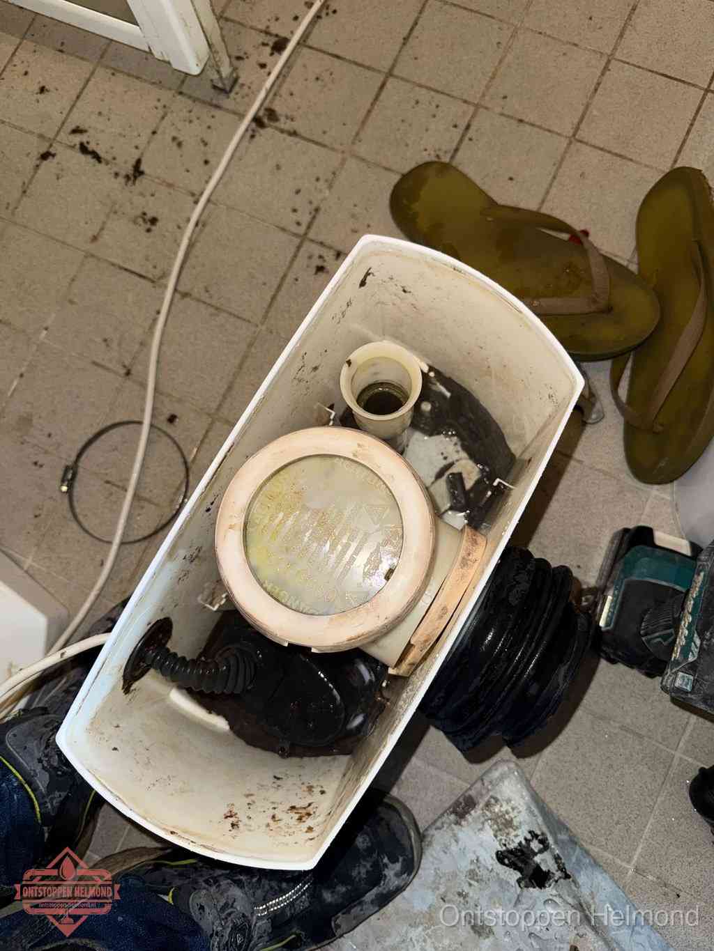 Een gedemonteerde toiletpot op de vloer met ontstoppingsveer en gereedschap ernaast, het verwijderen van een vastzittend voorwerp uit de wc in Helmond