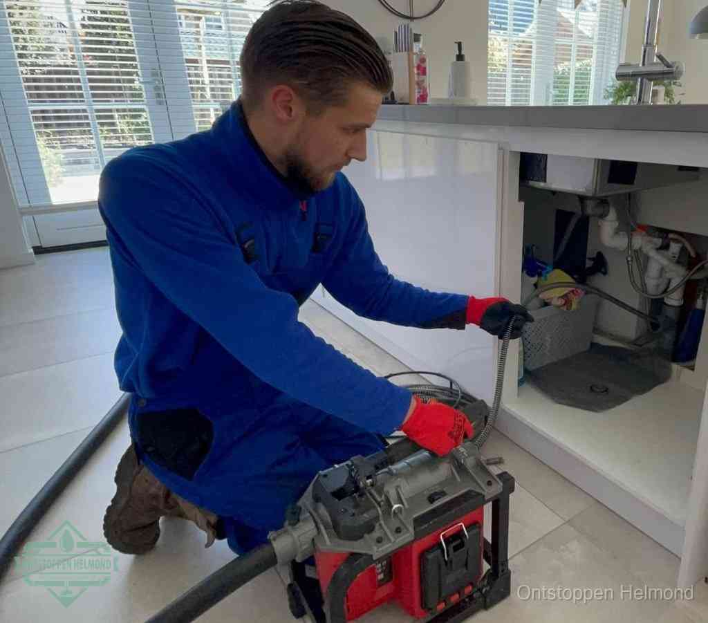 Een monteur met rode handschoenen knielt in een keuken en voert een elektrische ontstoppingsveer in tijdens het professioneel afvoer ontstoppen in Helmond